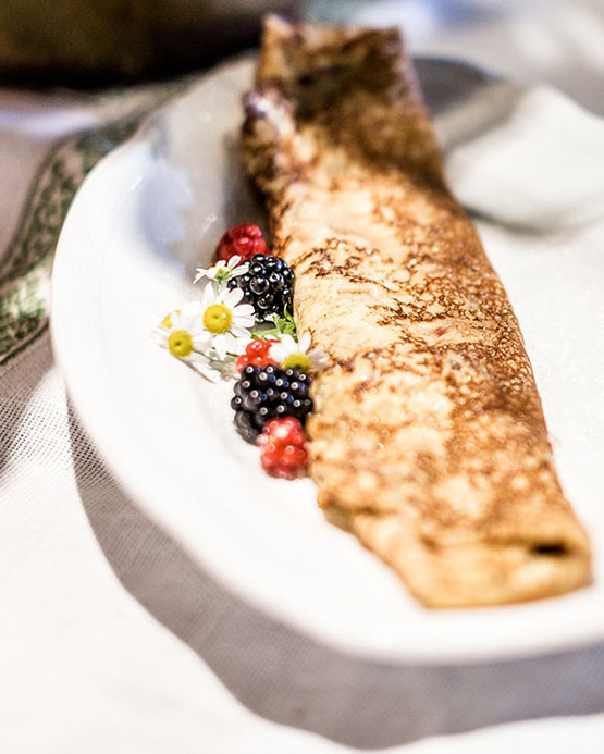 A rolled golden-brown crepe on a white plate, garnished with fresh blackberries, raspberries, and small white flowers.