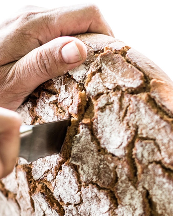 Hands slicing into a round loaf of rustic, crusty bread with a knife.