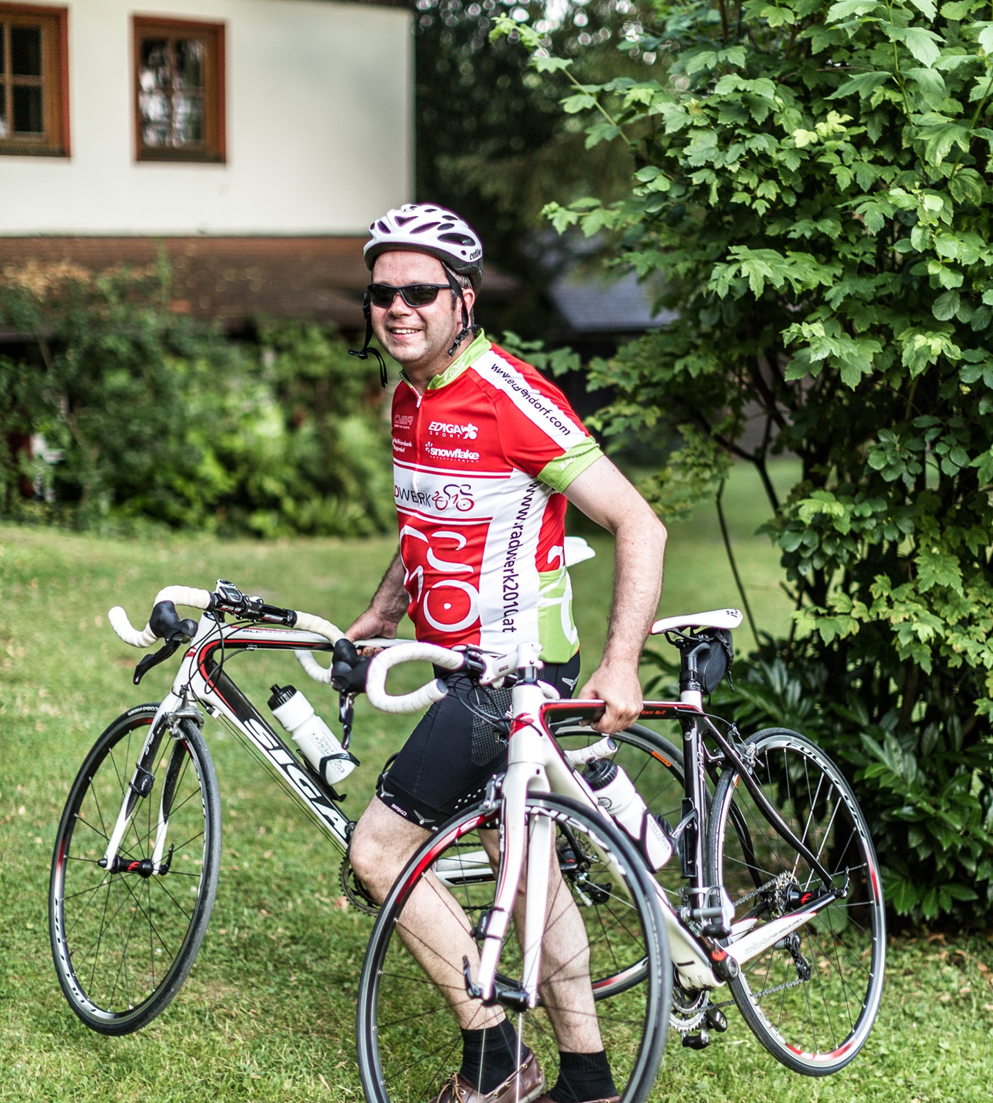 Smiling cyclist in a red and white jersey stands on grass, holding two white road bikes.