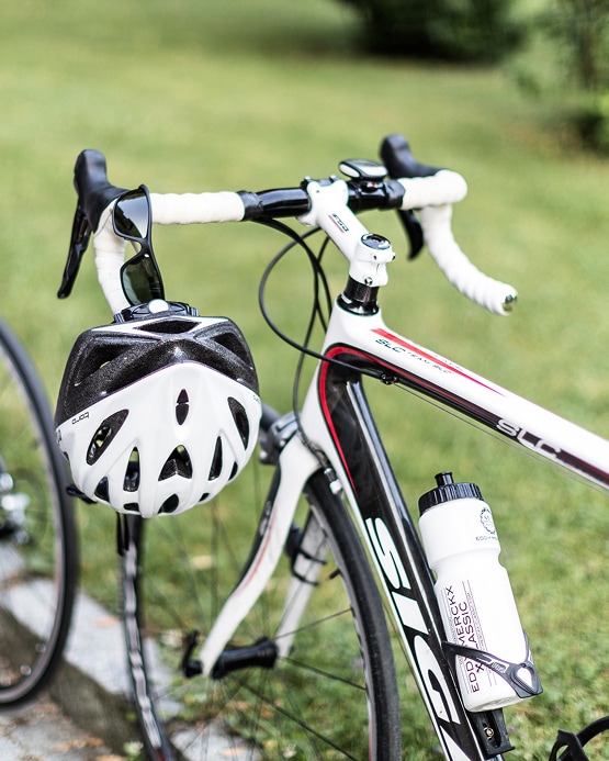 Close-up of a white and black road bike with a helmet and sunglasses hanging from the white handlebar tape.