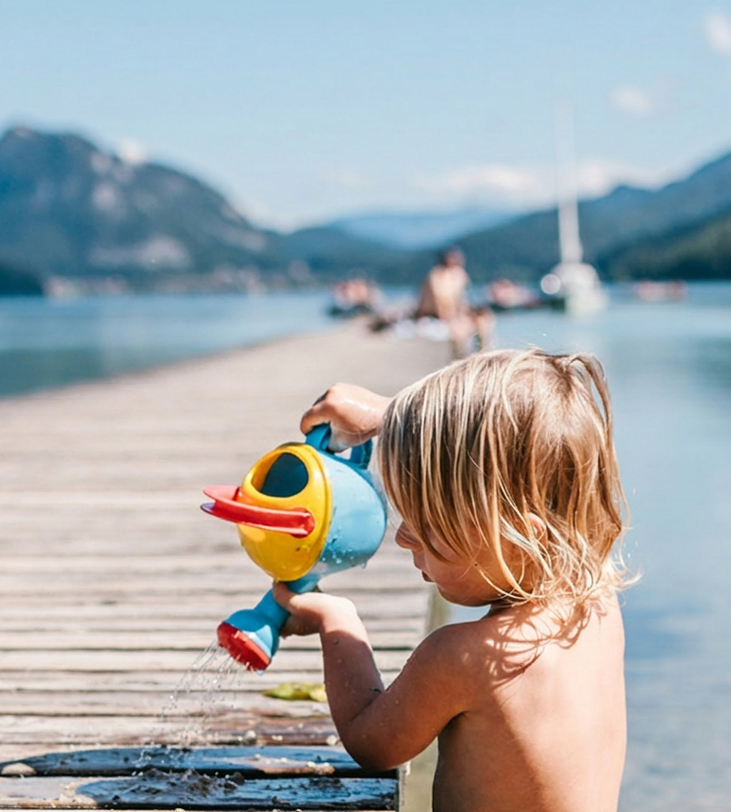 A young blonde child plays with a colorful watering can on a wooden pier by a scenic mountain lake.