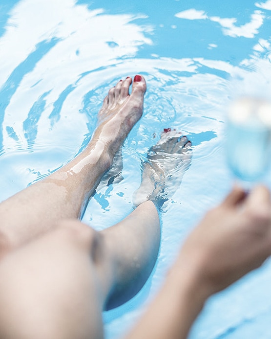 Close-up of a person's legs and feet with red nail polish dipping in a clear blue swimming pool.