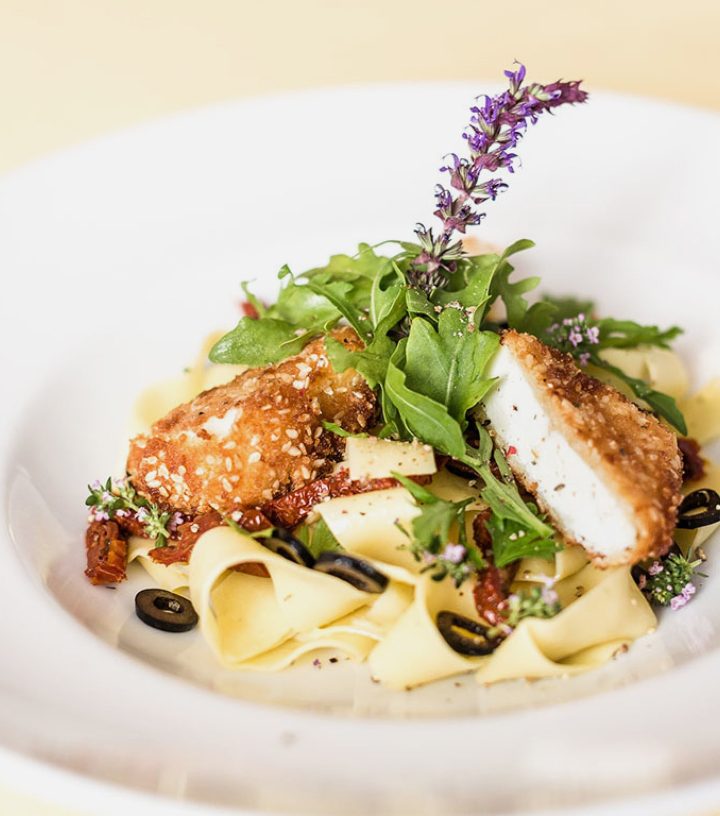 Plated pasta dish topped with sesame-crusted fried cheese, fresh arugula, black olives, and purple edible flowers.