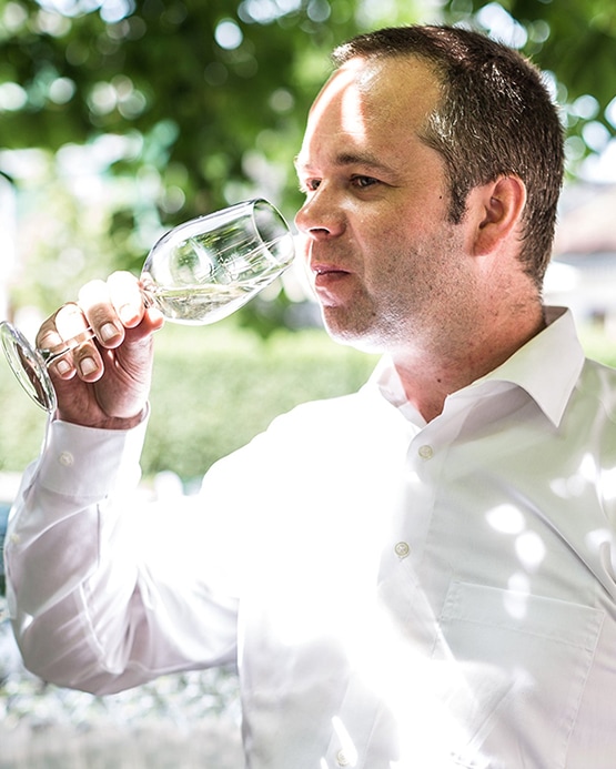 A man in a white shirt holding a wine glass to his lips, tasting white wine outdoors.