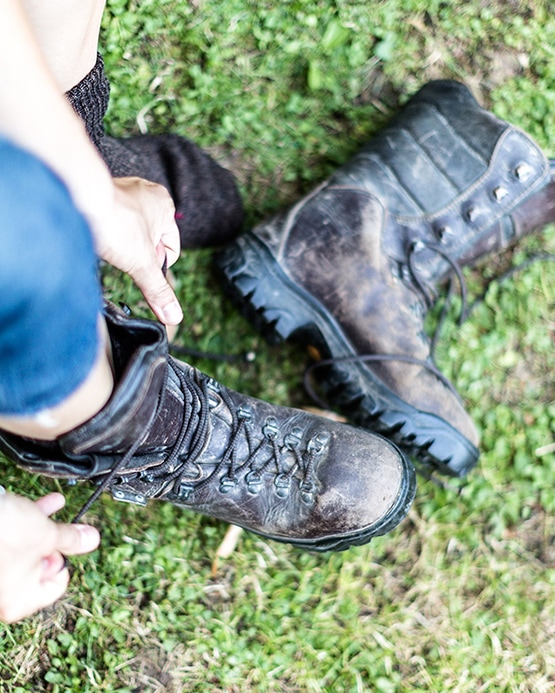 Overhead view of a person tying the laces of a rugged, dark leather hiking boot while sitting on green grass.