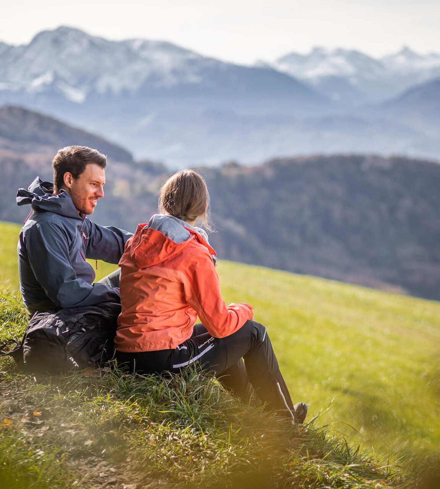 A man and woman in hiking gear sit on a grassy hillside, looking out at snowy mountains in the distance.