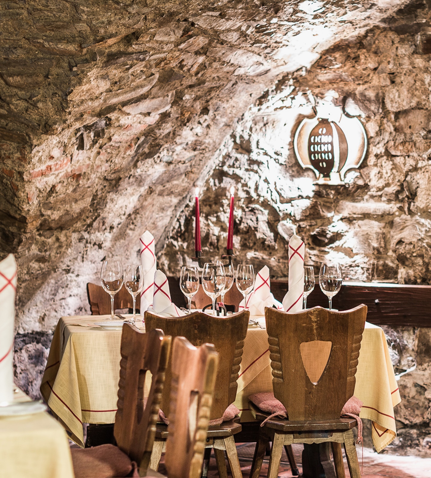 Dining table set with wine glasses and red candles in a rustic stone cellar with wooden chairs.
