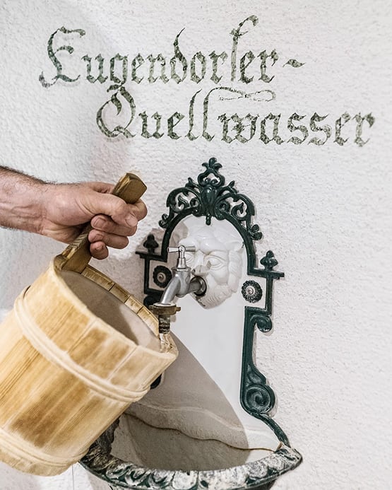 Ein Holzeimer wird unter einen Wandbrunnen gehalten. Darüber steht „Eugendorfer-Quellwasser“.