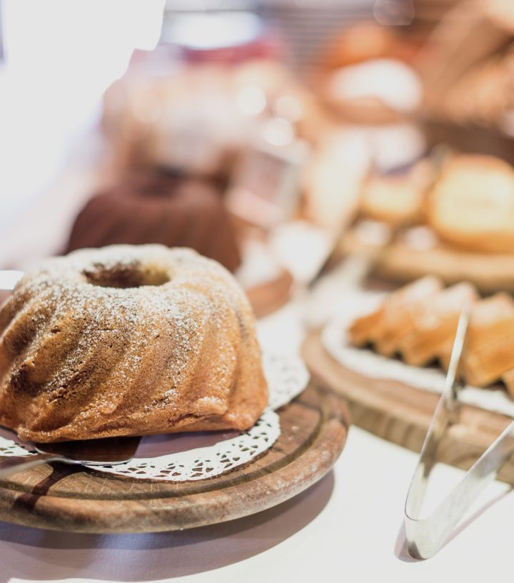 A bundt cake with powdered sugar sits on a wooden stand, surrounded by other pastries on a buffet table.