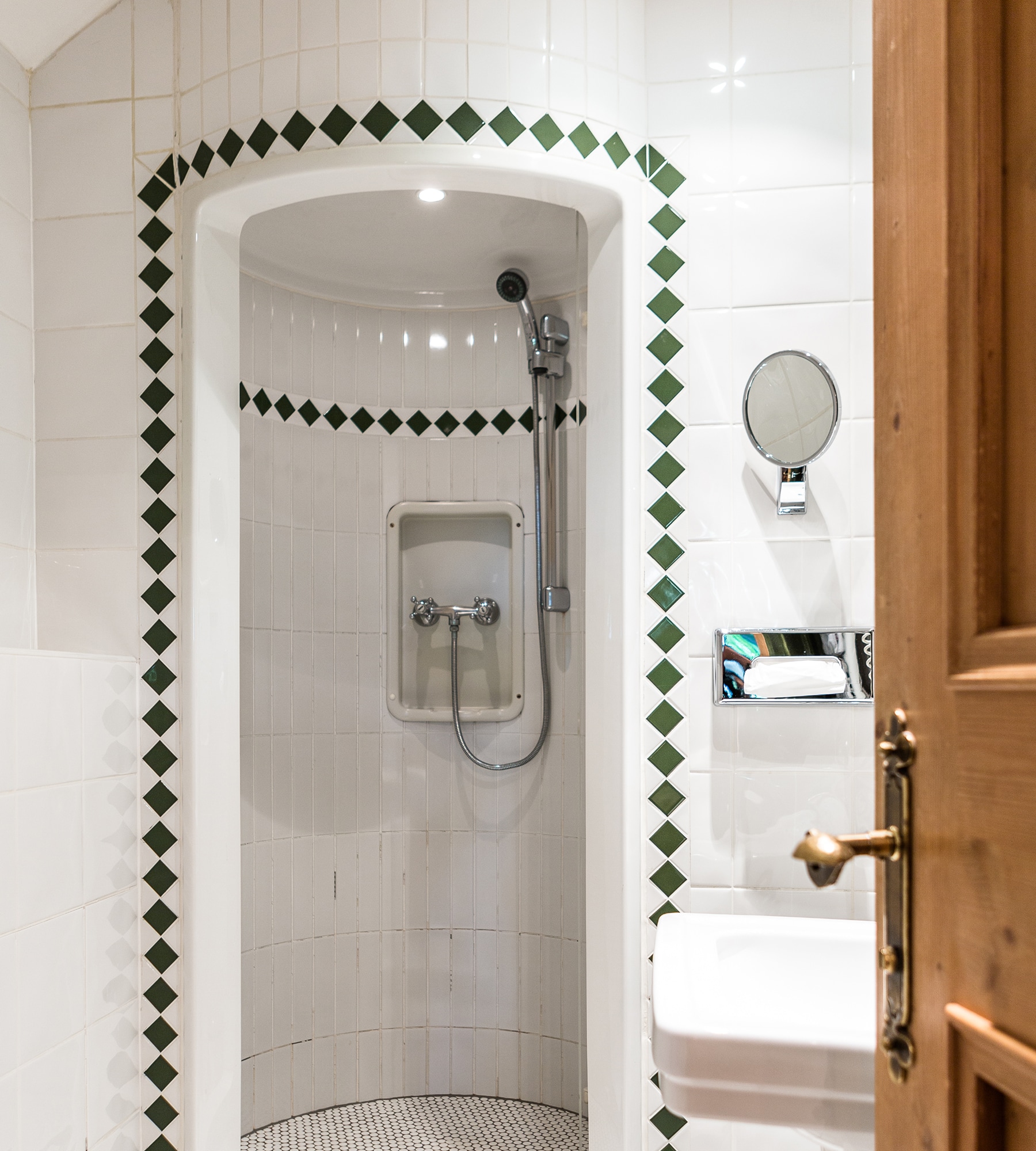 Tiled shower stall with green diamond border, viewed past an open wooden door next to a sink.