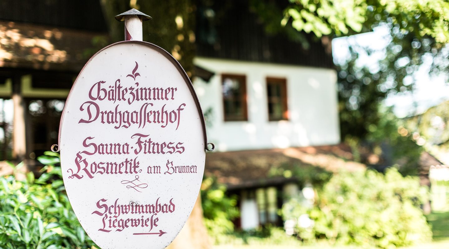 Oval sign for "Gästezimmer Drahgassenhof" in front of a white guesthouse with dark wood trim.
