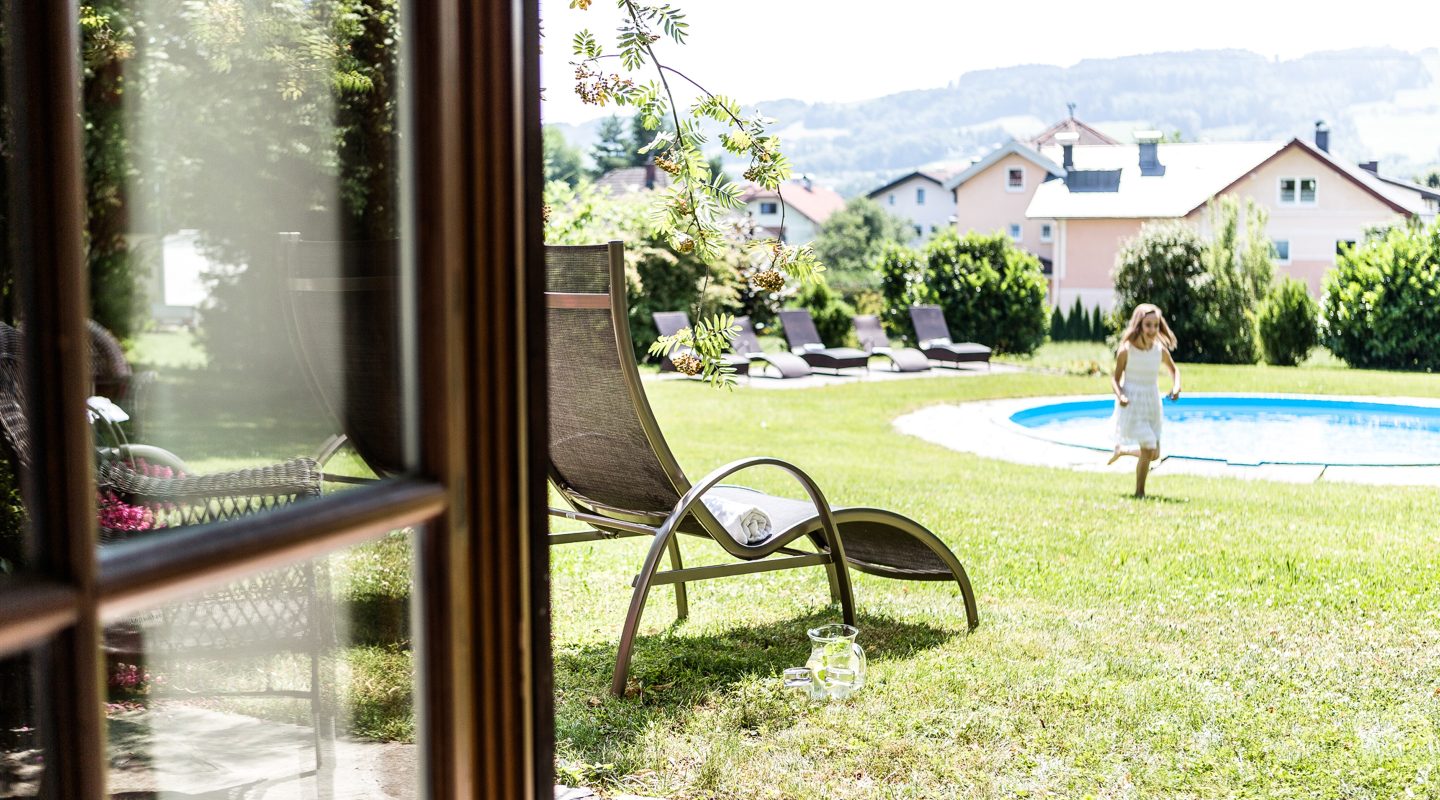 A girl in a white dress runs on a sunny lawn near a pool, seen from a window with lounge chairs nearby.