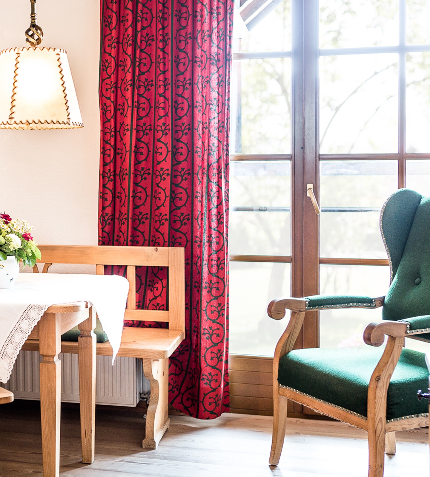 Cozy dining area with wooden bench, red patterned curtains, and a green upholstered armchair by a bright window.