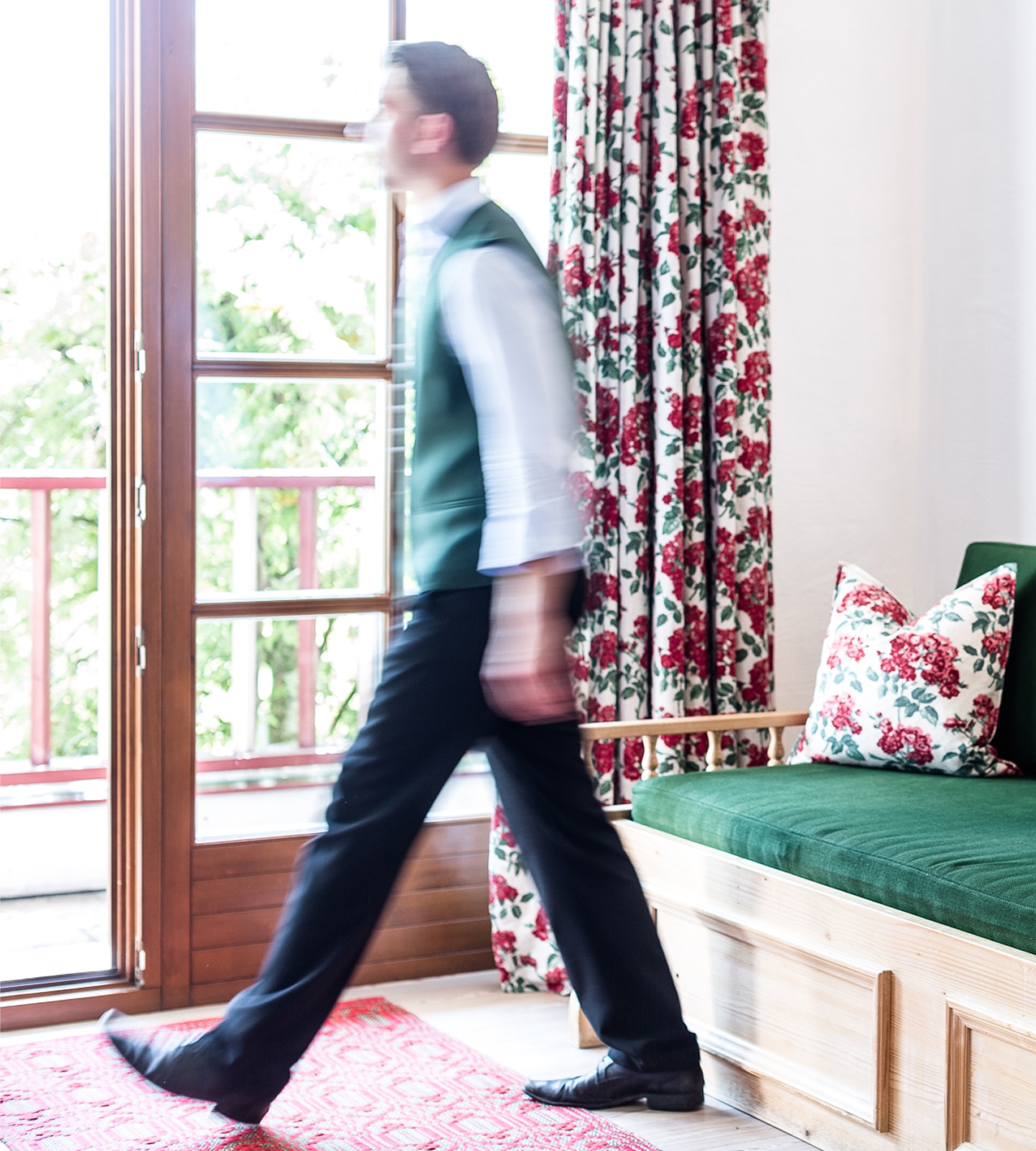 A motion-blurred man in a green vest and black trousers walks past a green wooden bench with floral cushions.