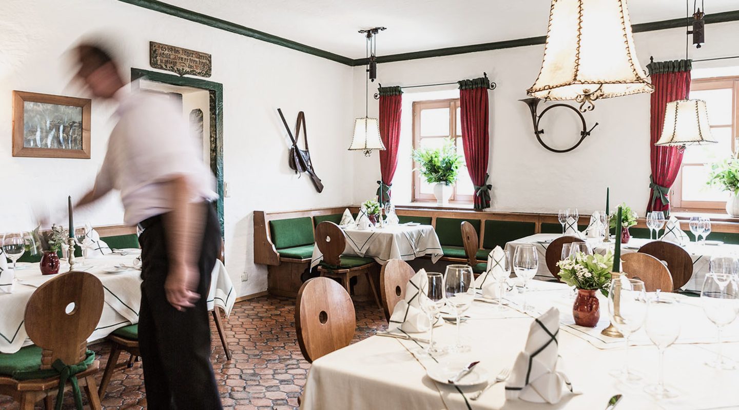 Rustic dining room with set tables, wooden chairs, red curtains, and a blurred figure of a waiter walking.