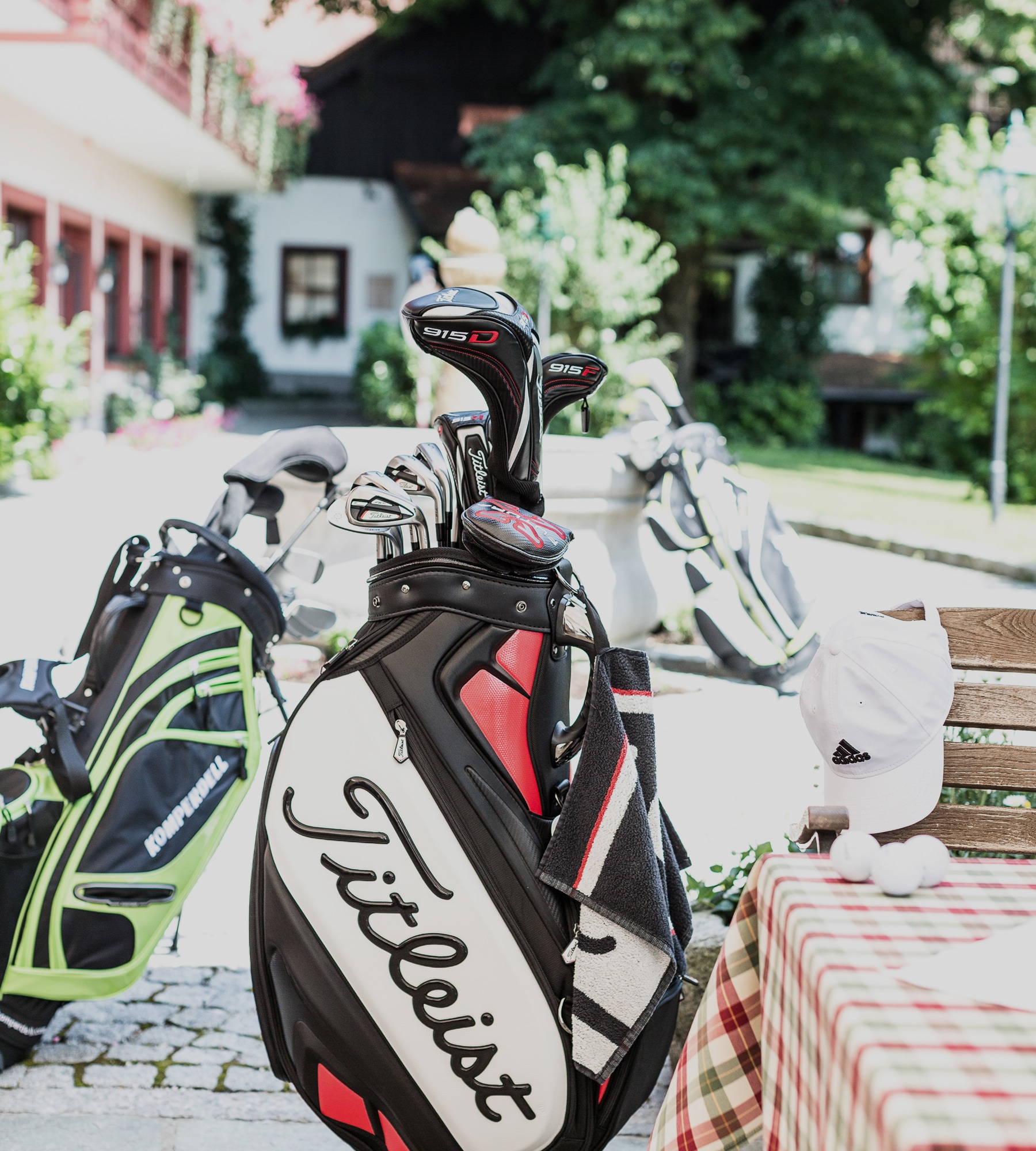 A Titleist golf bag filled with clubs stands outdoors near other golf bags, a table, and a white Adidas hat.