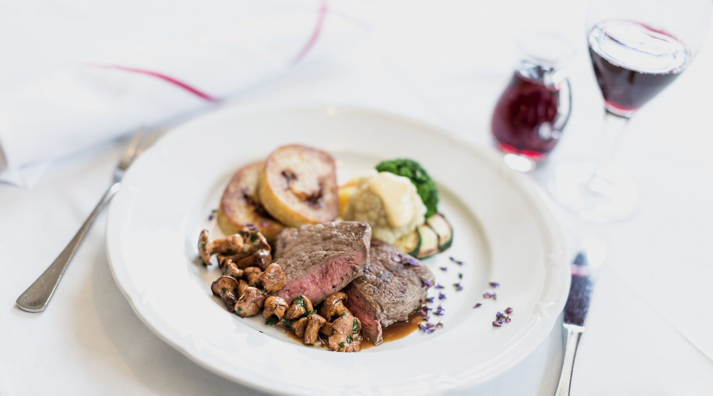 Ein Teller mit rosa gebratenem Rindersteak, Pfifferlingen, Knödeln, Gemüse und einem Glas Rotwein.