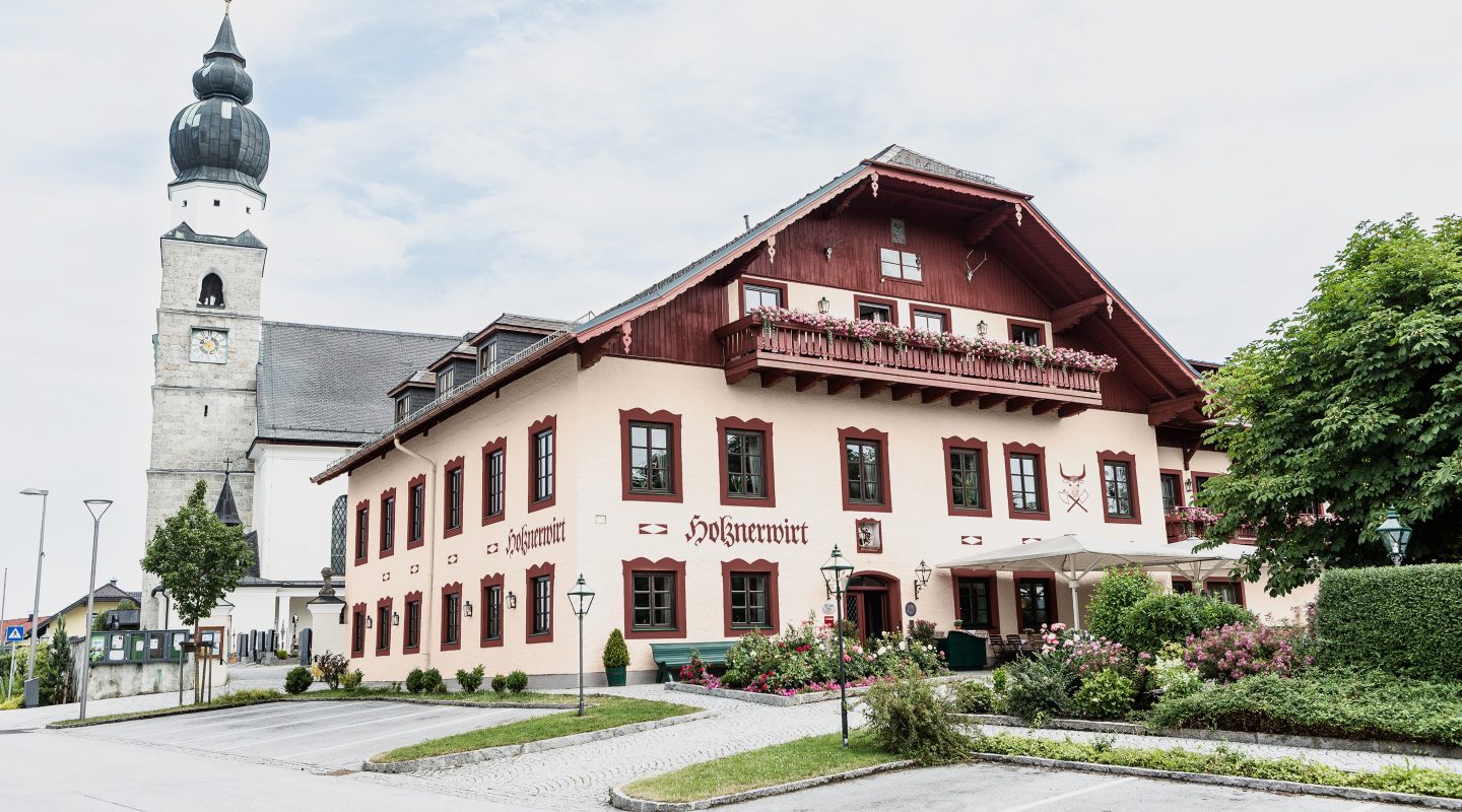 Der Gasthof Holznerwirt in Eugendorf mit dem Kirchturm im Hintergrund an einem bewölkten Tag.