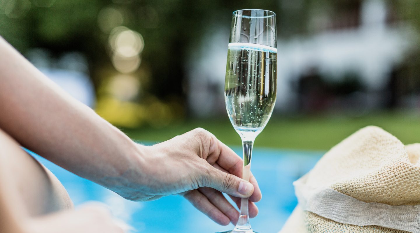Eine Hand greift nach einem Sektglas am Pool, daneben liegt ein Strohhut.