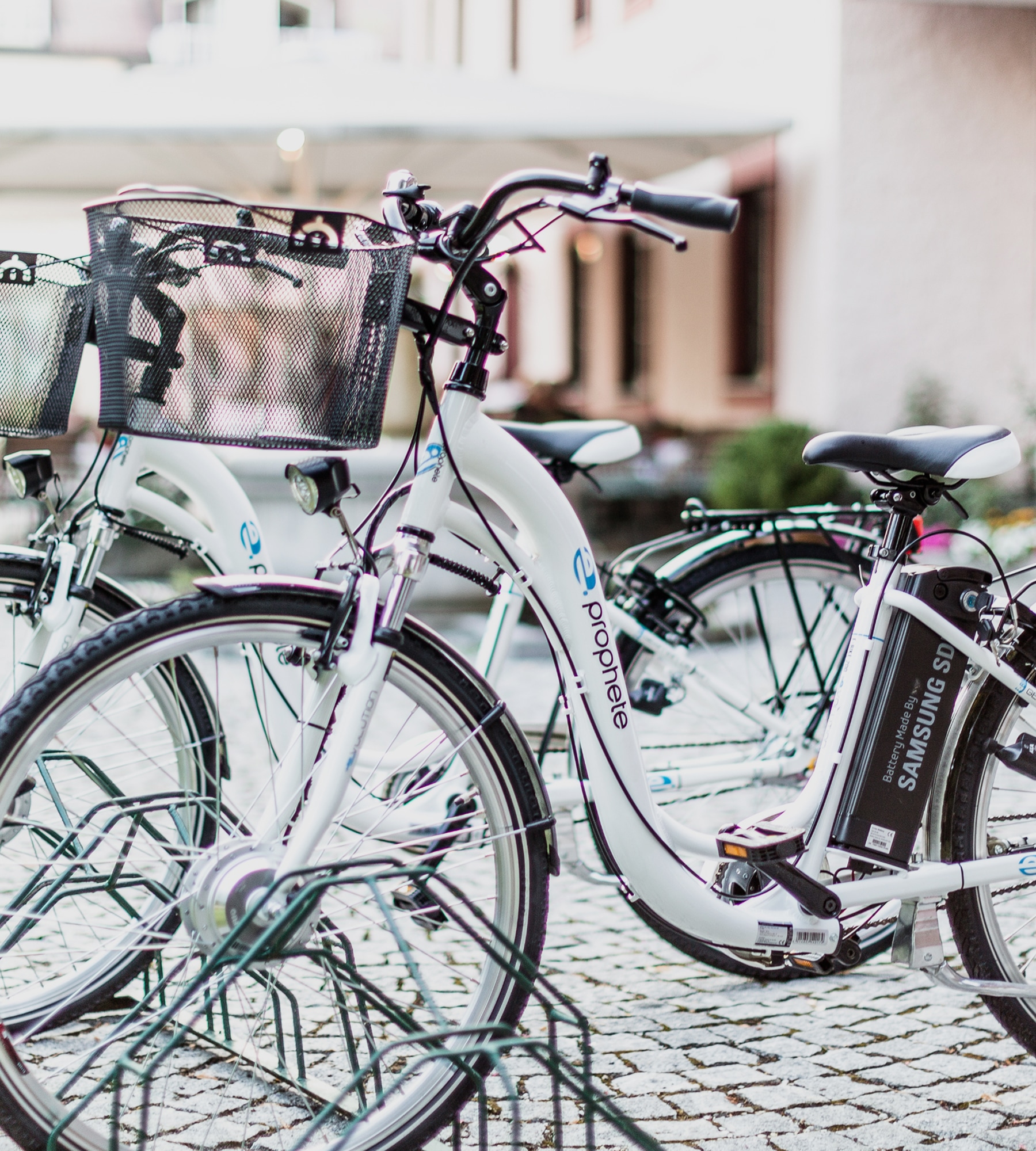 Weißes Prophete-E-Bike mit Samsung-Akku und Korb im Fahrradständer auf Kopfsteinpflaster.