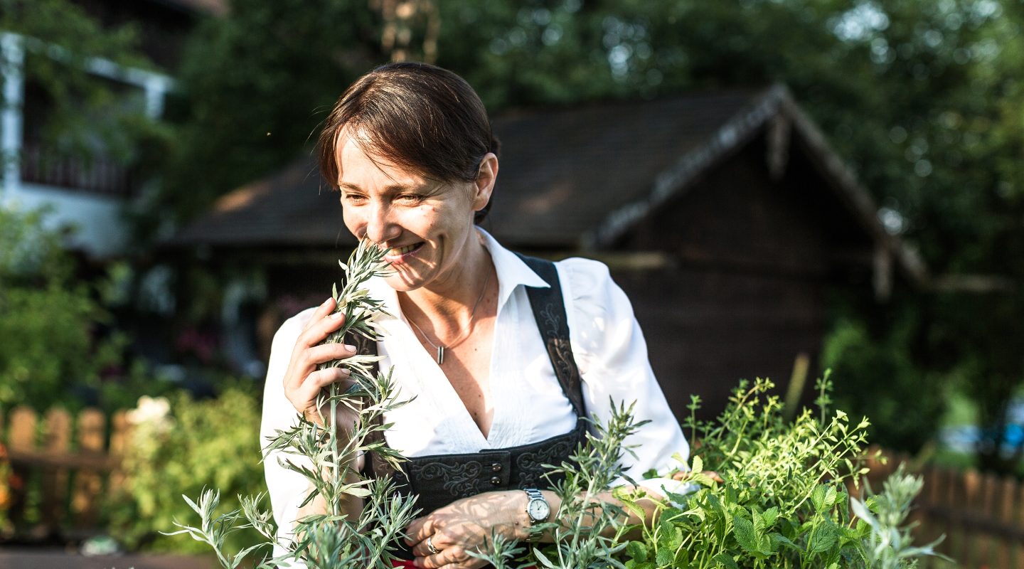 Smiling woman in traditional Bavarian dirndl smelling fresh herbs in an outdoor garden.