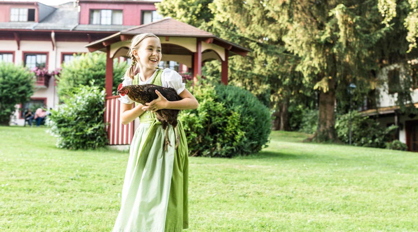 A smiling girl in a green traditional dirndl dress cradles a brown chicken in her arms on a grassy lawn.
