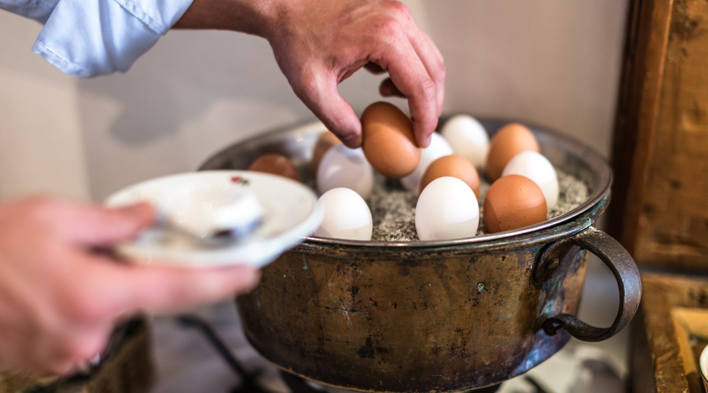 Close-up of a person's hand picking a brown egg from a rustic pot filled with sand and various eggs.