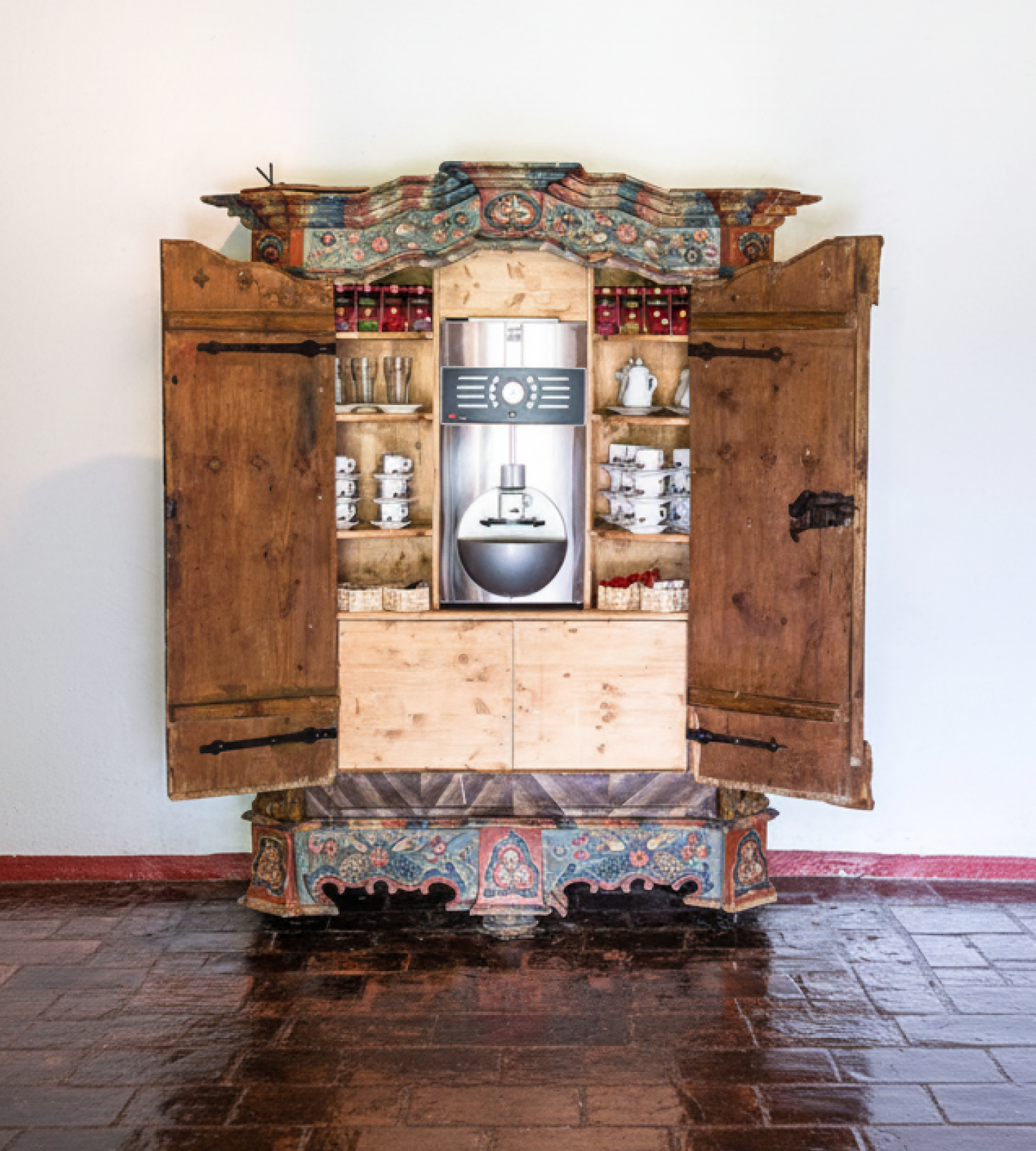 An open antique wooden cabinet with painted details, housing a modern coffee machine, cups, and glasses.