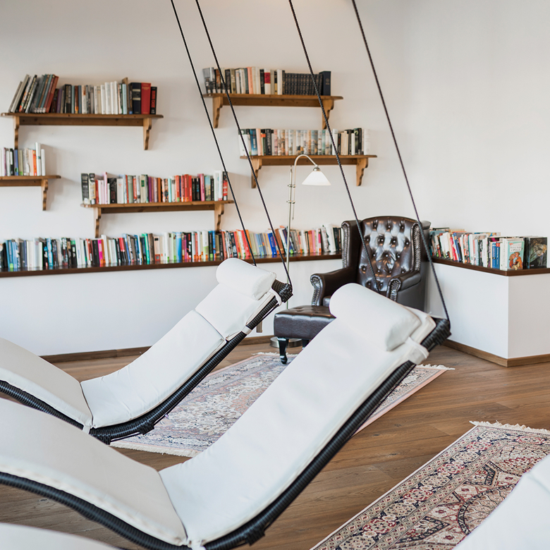 Modern library with hanging white loungers, wooden shelves filled with books, and a brown leather armchair.