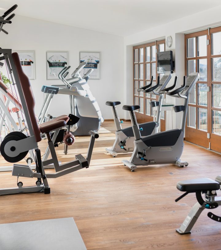 Bright gym with wooden floors, stationary bikes, a treadmill, and weight machines by glass doors.