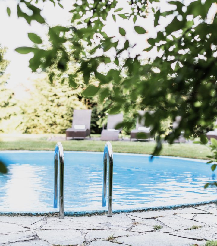An outdoor swimming pool with blue water and metal handrails, framed by green leaves with lounge chairs in the background.